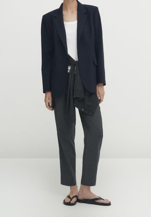 Personne portant un blazer bleu marine sur une chemise blanche avec un pull sombre noué autour de la taille, un pantalon gris foncé et des tongs noires.