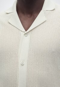 Άτομο με σκουρόχρωμο δέρμα που φορά ένα κρεμ πουλόβερ με υφή πλεκτού, με μυτερό γιακά και δύο ορατά κουμπιά.