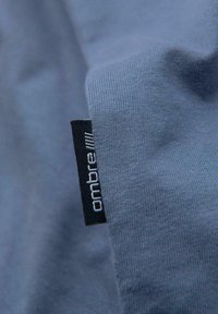 Tissu bleu avec une étiquette de vêtement noire portant le nom de la marque "ombre", cousue dans la couture du textile.