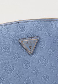 Μπλε τσάντα από δερματίνη με ανάγλυφα σχέδια, επιχρυσωμένη τρίγωνη πλακέτα με λογότυπο και φερμουάρ στην κορυφή.