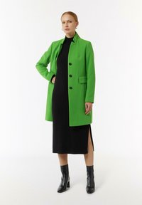 Helle grüne Jacke mit Reverskragen, vier schwarzen Knöpfen und seitlichen Taschen, getragen über einem schwarzen Kleid mit seitlichem Schlitz und schwarzen Stiefeln.