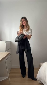 Donna sorridente all'interno, indossa una maglietta bianca e pantaloni neri, tiene una giacca scura, in piedi vicino a una scrivania e a un letto in una stanza minimalista.