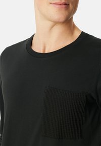 Zwart shirt met lange mouwen, featuring een textuur pocket, ronde hals en een gladde stof. De pocket heeft een geribbeld patroon voor extra detail.
