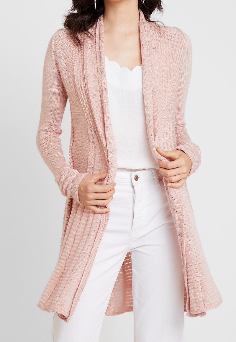 Ljusrosa stickad cardigan med ribbad textur, öppen framsida, långa ärmar och avslappnad passform, i kombination med en vit topp och jeans.