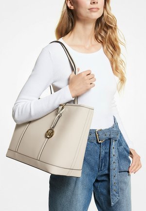 Frau trägt weißes Langarmshirt und blaue Jeans mit Gürtel, trägt eine beige Leder-Umhängetasche mit goldenem Emblem auf der Schulter.