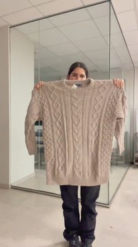 Une personne tient un pull beige à torsades devant son torse dans une pièce moderne avec des murs en verre et un plafond carrelé.