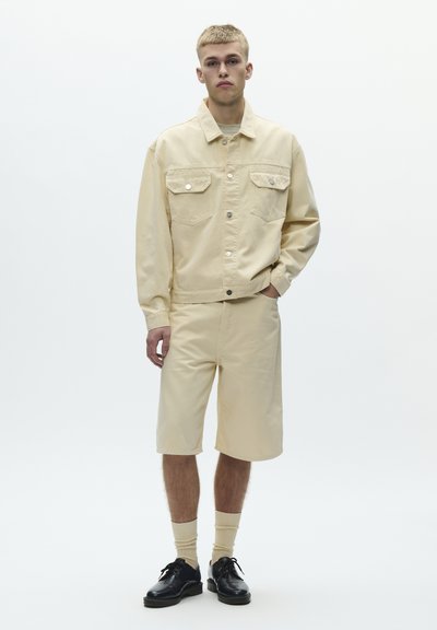 Young man wearing beige jacket and matching knee-length shorts, beige socks, and black lace-up shoes, standing against a plain background.