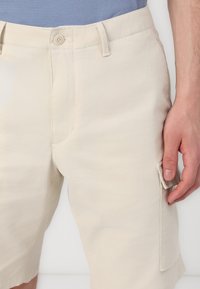 Pantaloni scurți din bumbac bej, cu închidere prin buton, buzunare laterale și un tiv texturat, prezentând un finisaj de țesătură netedă și moale.