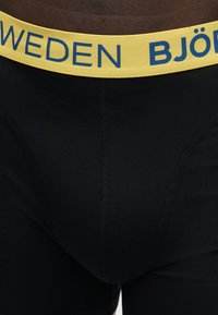 Μαύρα αντρικά εσώρουχα boxer με ορατή ραφή μπροστά και κίτρινη μέση με μπλε γράμματα που γράφουν "SWEDEN BJÖR."