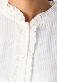 Nærbillede af en hvid ribbet stoftop med flæser langs knapstolpen og halsudskæringen, placeret på en hudfarvet baggrund.