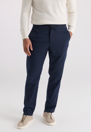 Uomo con pantaloni blu navy con coulisse, scarpe da ginnastica beige e camicia bianca a maniche lunghe, in piedi con le mani parzialmente nelle tasche.