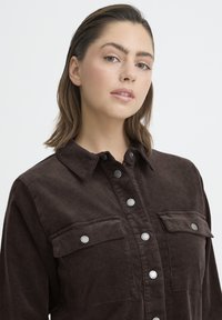 Mlada žena svijetle puti i smeđe kose, odjevena u tamnosmeđu kordurovu košulju s gumbima i džepovima s klapnama, gleda blago u stranu.