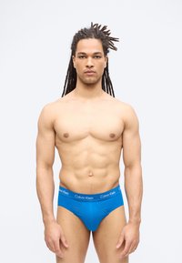 Izmos férfi, hosszú dreadlockokkal, előre néz, kék Calvin Klein alsónadrágot visel, egy sima világos háttér előtt.