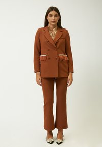 Blazer marrone doppiopetto con dettagli in piume e gioielli sulle tasche, abbinato a pantaloni marroni a zampa d'elefante e scarpe con decorazioni.