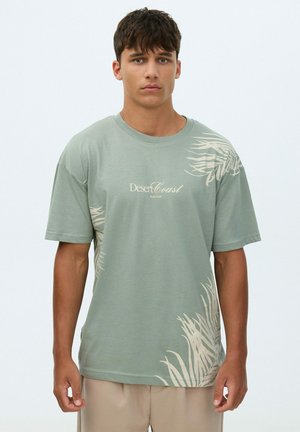Joven con una camiseta verde claro con estampados de hojas tropicales beige y texto "Desert Coast", combinado con pantalones beige, de pie sobre un fondo blanco.