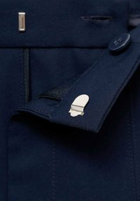 Navystoffdetails mit einem silbernen Clip, einem Knopf mit Logo und einem Reißverschluss. Die Textur ist glatt mit sauberer Naht und taillierten Kanten.