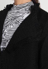 Cardigan noir texturé avec des détails en frange, superposé à un haut blanc à col montant présentant des rayures abstraites noires. Vue rapprochée des tissus.