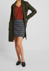 Manteau en laine vert olive, haut à col en v rouille, mini-jupe en tweed multicolore à base bleue et chaussures plates en cuir noir.