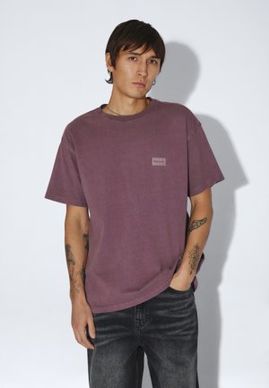 Ung mand med kort mørkt hår iført en falmet lilla t-shirt og sorte jeans, står med den ene hånd i lommen foran en ensfarvet baggrund.