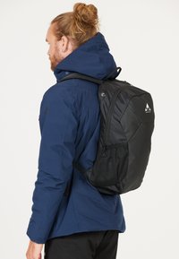 Svart ryggsäck med texturerad tyg, justerbara remmar och sidoficka i mesh. Bärs över en blå jacka och visar upp en strömlinjeformad design.
