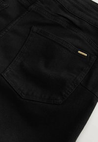Poche arrière d'un jean en denim noir avec une petite plaque métallique rectangulaire gravée du mot « NEXT » dans le coin supérieur droit.