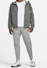 Homme portant un sweat à capuche gris zippé, un t-shirt blanc, un pantalon de jogging gris et des baskets blanches avec des touches noires, debout devant un fond blanc.