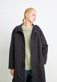 Svart lang regnjakke med hette, med en front glidelås og trykknapp lukking. Brukt over en lys grønn hettegenser og blå jeans.