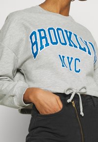 Felpa grigia con testo blu "BROOKLYN NYC". Presenta un collo rotondo, maniche lunghe e un cordino all'orlo. Indossata con pantaloni neri.