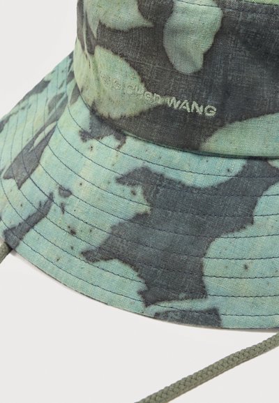 Πράσινο και μαύρο καμουφλάζ καπέλο τύπου bucket με κεντημένο λογότυπο "alexander WANG" και πλεγμένο λουράκι στο πηγούνι σε λευκό φόντο.