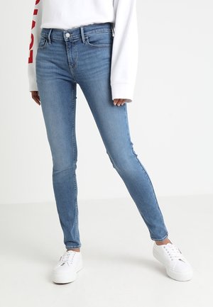 Personne portant un jean skinny bleu clair associé à des baskets blanches et un haut blanc à manches longues, debout les jambes légèrement écartées.