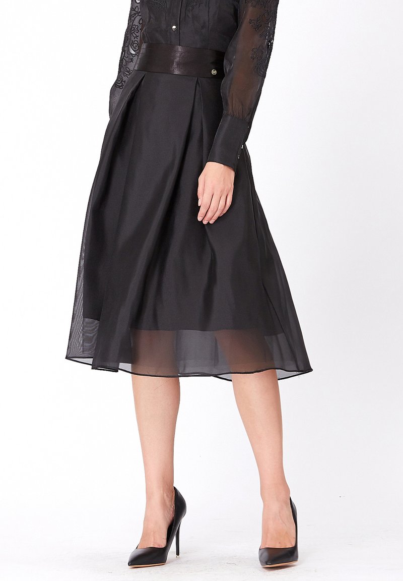 Jupe midi noire en chiffon superposé avec une taille cintrée en satin, présentant des plis et un ourlet transparent. Associée à des chaussures noires à talons hauts.