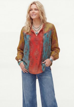 Femme portant un chemisier à motifs rouge, bleu et moutarde à manches longues, un jean bleu, des bijoux turquoise et plusieurs bracelets en argent.