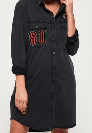 Femme portant une robe en denim noire boutonnée avec des manches retroussées et des patchs brodés rouges et dorés.