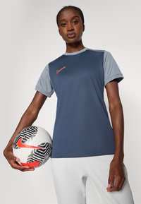 Tummansininen ja harmaa lyhythihainen urheilupaita, jossa on Nike-logo; pitää mustaa ja valkoista jalkapalloa, jossa on punaisia yksityiskohtia ja geometrisia kuvioita.