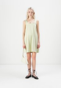 Vestido sem mangas em padrão xadrez verde claro, combinado com uma mala branca e sapatos planos pretos com atacadores. Design simples, ajuste descontraído, até ao joelho.