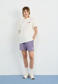 T-shirt à manches courtes blanc cassé avec logo rouge, associé à un short en velours côtelé violet clair avec un design graphique blanc. Chaussettes blanches et baskets.