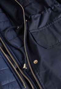 Marineblå jakke med quiltet indre, guld lynlås, snoretræk og en patch-lomme. Glat tekstur med et strømlinet design.