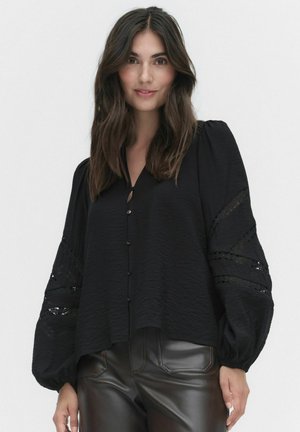 Zwarte blouse van gestructureerde stof, lange puffmouwen, kantaccenten en knoopsluiting aan de voorkant. Gecombineerd met zwarte leren broeken.