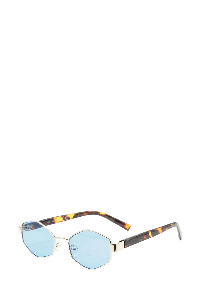 Occhiali da sole esagonali con lenti azzurro chiaro, montatura in metallo dorato e bracci con motivo tartaruga poggiati su una superficie bianca.