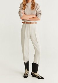 Beiger Pullover, hellgraue, gefaltete Hose, brauner Gürtel und schwarze, mit Schlangenhautmuster verzierte Cowboy-Stiefel mit spitzer Zehenform.