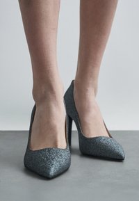 Pelēki mirdzoši augstpapēžu zābaki ar norād�ītu purngalu un gludu tekstūru. Apaviem ir zema griezuma vampi un slaidi papēži.