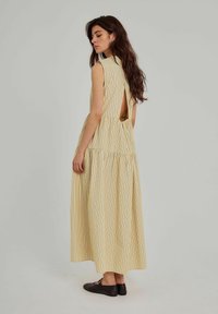 Gulstribet maxikjole med åben ryg, ærmeløst design og en svajet skirt. Sorte flade sko fuldender outfittet.