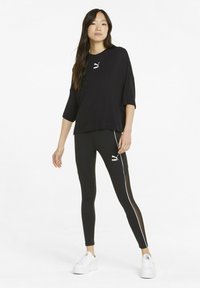 Mustat urheilupaidat, joissa on lyhyet hihat ja Puma-logo, yhdistetty mustiin legginsseihin, joissa on sivustripit ja läpinäkyvä paneeli, käytössä valkoisten lenkkareiden kanssa.