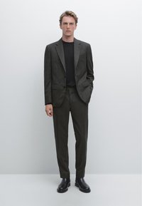 Abito su misura grigio scuro con rever a lancia, abbinato a una camicia nera e stivali in pelle nera. Tessuto liscio, taglio slim, design minimalista.