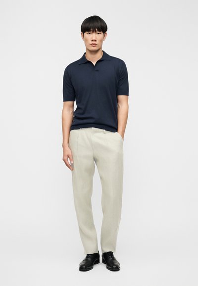 Polo de manga corta de color azul marino con cuello, combinado con pantalones beige claro y zapatos negros, posando frente a un fondo sencillo.