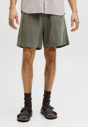 Mężczyzna w oliwkowo-zielonych szortach, ciemnych skarpetach i czarnych sandałach z klamrą stoi na jednolitym białym tle.