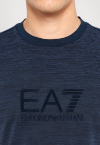 Tamsiai mėlyna marškinėliai su apvaliu iškirpimu, pagaminti iš tekstūruotos medžiagos. Pavaizduotas didelis tamsus "EA7 EMPORIO ARMANI" logotipo atspaudas ant krūtinės.