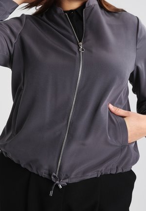 Kobieta ubrana w szarą kurtkę na zamek z regulowanym ściągaczem na dole, z jedną ręką w kieszeni, widoczna od szyi do bioder.