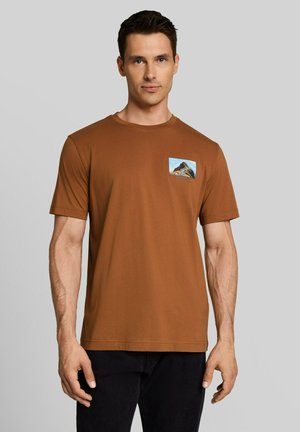 Man wearing a brown short-sleeve t-shirt with a small mountain graphic on the left chest against a plain gray background.