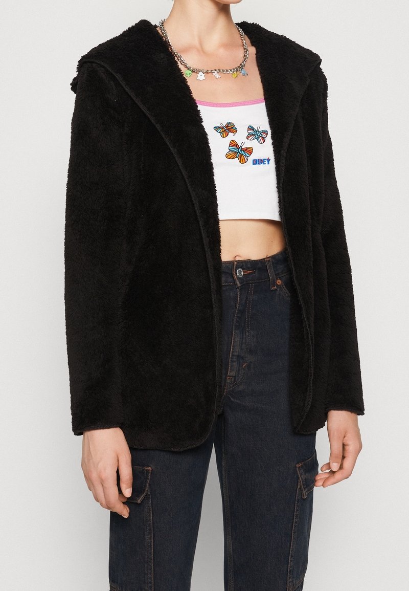 Zwarte, fuzzy stoffen jas met een sjawl-kraag en een open voorkant ontwerp. Heeft lange mouwen en een ontspannen pasvorm. Draagt over een cropped top.